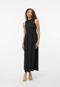 Robe maxi noire sans manches avec un buste froncé, une jupe fluide, une texture douce et un col montant. Associée à des chaussures à talons ouverts jaunes.