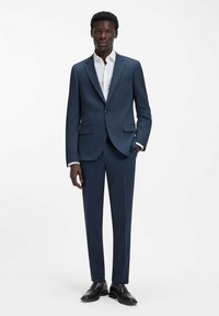 Abito blu navy con tessuto strutturato, vestibilità sartoriale, giacca monopetto con due bottoni, ampi revers, abbinato a una camicia bianca e scarpe nere.