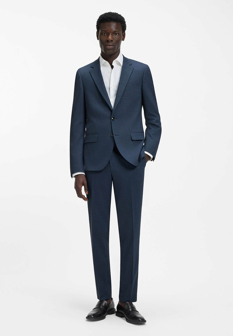 Abito blu navy con tessuto strutturato, vestibilità sartoriale, giacca monopetto con due bottoni, ampi revers, abbinato a una camicia bianca e scarpe nere.