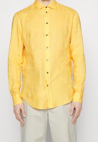 Žlutá lněná košile s dlouhými rukávy, špičatým límcem a jemnou strukturou. V kombinaci se světle zbarvenými kalhotami.