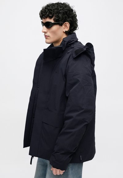 Tumši zila jaka ar augstu apkakli, rāvējslēdzēju un spiedpogām, teksturētām panelēm un vairākām kabatām. Raksturo glīts, mūsdienīgs dizains.