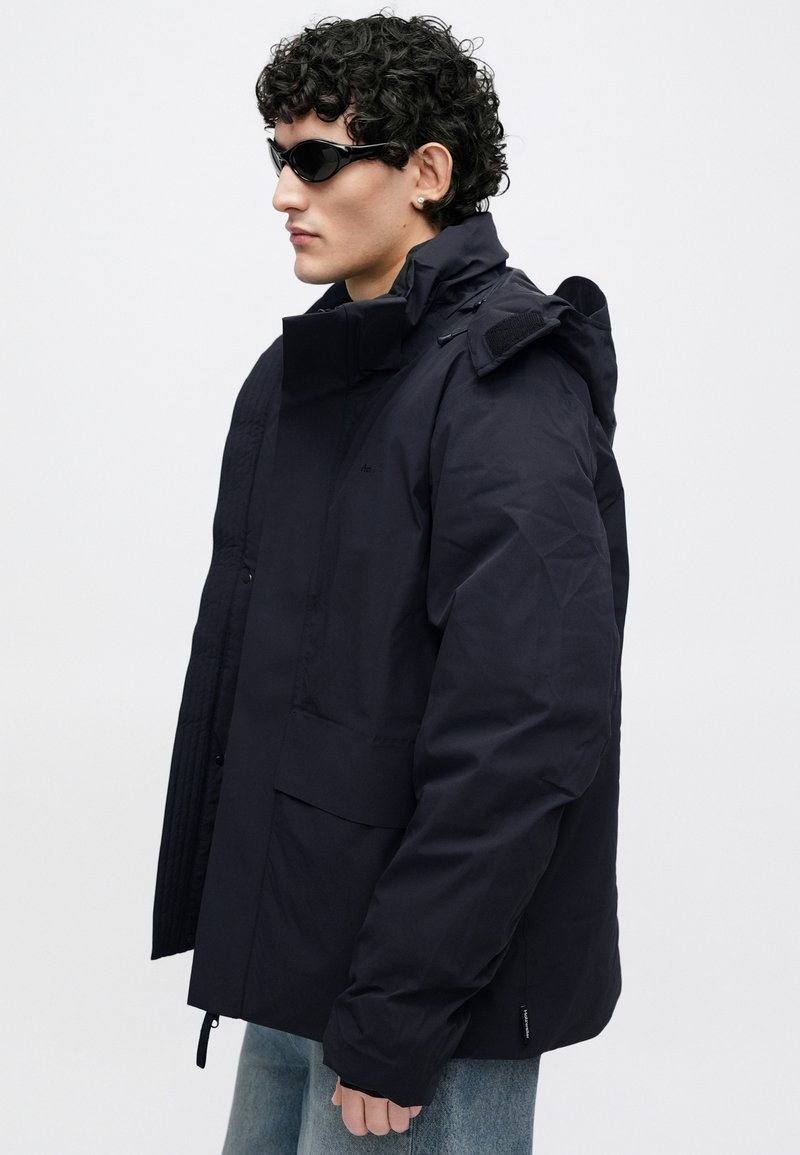 Navy blue jacket with a high collar, zip and snap closures, textured panels, and multiple pockets. Features a sleek, modern design.