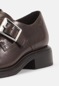 Brown leather shoe with a rectangular silver buckle and stitched detailing, featuring a block heel and a smooth textured surface.