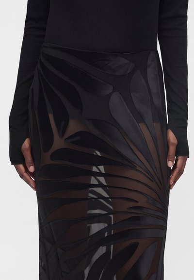 Personne portant un haut noir ajusté et une jupe noire transparente avec de grands motifs abstraits de feuilles en velours. Les mains reposent le long du corps.