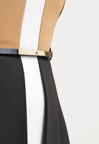 Robe bicolore présentant des sections beige, blanche et noire, en tissu lisse, avec une ceinture métallique accentuant la taille.