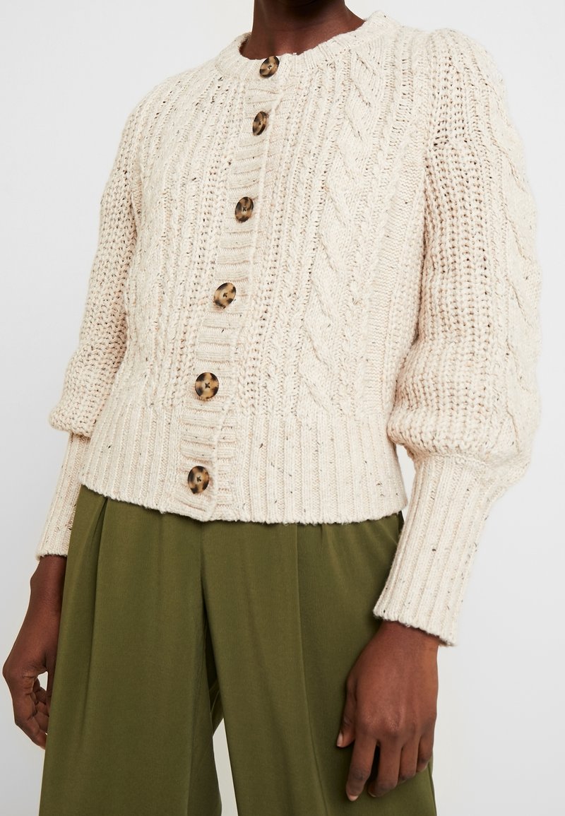 Personne portant un cardigan crème en maille torsadée boutonné avec des manches bouffantes et un pantalon large vert olive.