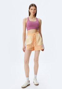 Sujetador deportivo en púrpura combinado con pantalones cortos de un naranja claro que tienen bolsillos amarillos y lavanda. La modelo lleva zapatillas y calcetines blancos.