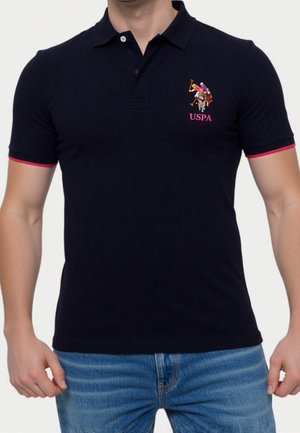 Homme portant un polo noir USPA à manches courtes avec bordure rouge, associé à un jean bleu.