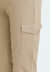Beige cargobukser med en glat overflade, der har en sidelomme med låg og syninger på stoffet.