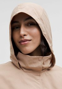 Femme portant une veste à capuche beige avec un col haut, affichant un léger sourire, devant un fond clair.