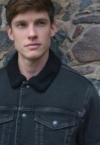 Melna džinsa jaka ar pūkainu melnu iekšu, kas aprīkota ar pogu aizdari un divām krūšu kabatām, pret akmens sienas fona.