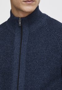 Suéter de punto azul marino con cierre de cremallera y cuello alto, que presenta un patrón texturizado y detalle en cremallera negra. Vista en primer plano de la tela.
