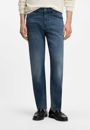 Mann trägt mittelblaue Straight-Leg-Jeans, cremefarbenen Pullover und schwarze Ledermokassins, steht vor weißem Hintergrund.