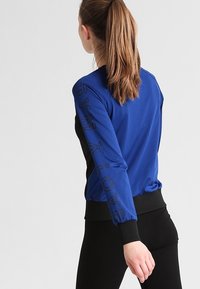 Giacca sportiva blu con accenti neri, maniche lunghe e lettering testurizzato sulla manica. Realizzata in tessuto traspirante con un design aderente.