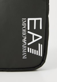 Sac noir arborant un texte blanc audacieux : "EA Emporio Armani" avec un logo stylisé, mettant en avant un design épuré et moderne.