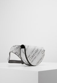 Calvin Klein-väska med vitt yttre, tryckt med svart logotekst, halvcirkelformad, grå rem och slät yta.
