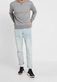 Šedá mikina s kulatým výstřihem, světlé modré džíny a černé sportovní boty. Mikina má žebrované manžety a lem. Celkově pohodlný outfit.