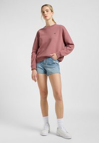 Sudadera rosa con cuello redondo, corte holgado; combinada con shorts de denim azul con bordes deshilachados y un parche floral. Zapatillas blancas completan el conjunto.