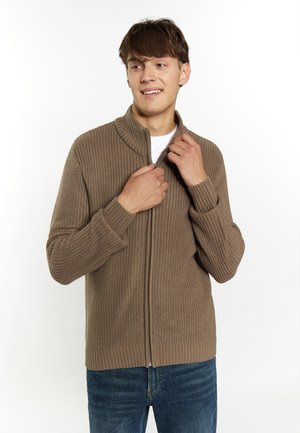 Cardigan tricotat maro cu un guler înalt, fermoar frontal, manșete cu striații și țesătură texturată. Purtați peste o cămașă albă și asortat cu blugi albaștri.