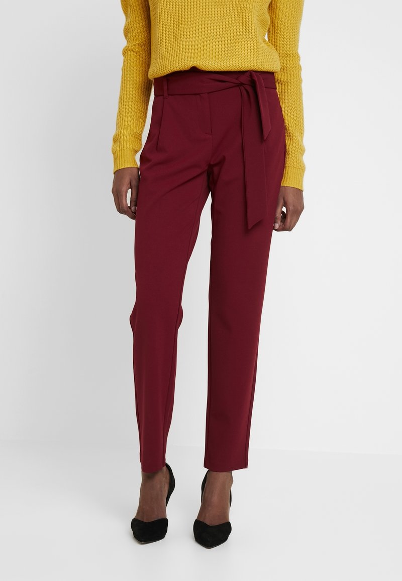 Pantalons bordeaux avec une ceinture à nouer, jambes fuselées et texture lisse, associés à un pull jaune texturé et des talons noirs.