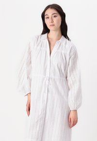 Rochie albă cu nasturi, cu mâneci lungi, având dungi verticale și detalii din dantelă. Talie ușor strânsă cu un cordon. Textură moale.