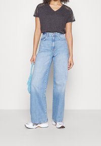 Camiseta gris de pico con encaje, vaqueros de tiro alto de pierna ancha azul claro, zapatillas blancas con detalles y un bolso de rafia azul.