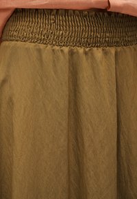 Κοντινή λήψη ενός φούστα ελιάς με ελαστική ζώνη με πιέτες και μια απαλή πορτοκαλί γαρνιτούρα που φαίνεται από πάνω.