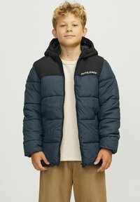 Pildīta jaka ar tumši zilu ķermeni un melnām plecu akcentiem, ar rāvējslēdzēju priekšpusē, kapuci un baltu logo uz krūts. Cieta, quilted dizaina.