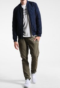 Marinblå kviltad bomberjacka över en grå T-shirt, kombinerad med olivgröna cargo-byxor och vita sneakers. Minimalistisk design, avslappnad passform.