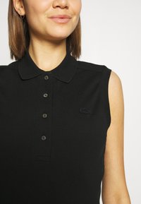 Čierna bezrukávová polo košeľa s golierom, ktorá má jemné gombíkové zapínanie a vyšívané logo v zladenej čiernej farbe na ľavej hrudi.