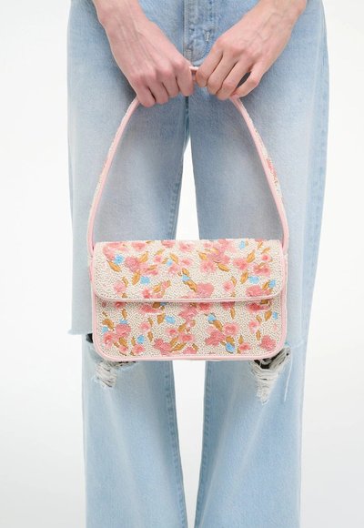 Personne tenant un sac à main rose floral perlé devant un jean bleu clair déchiré, les mains agrippant la poignée du sac.