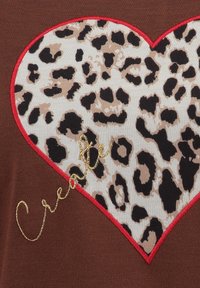 Hjerte-patch med leopardmønster og rød kant på brunt stof, med det broderede ord "Creative" i guld henover hjertet.