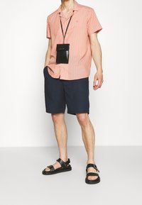 Person som bär en korall- och vitrandig kortärmad skjorta, marinblå shorts, svarta sandaler och en svart nyckelhållare med snöre, står mot en vit bakgrund.