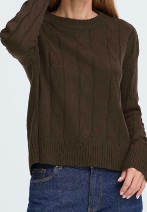 Kvinde iført en mørkebrun kabelstrikket sweater med ribkant forneden og ved ærmerne, kombineret med blå denimjeans, hånd hvilende nær taljen.