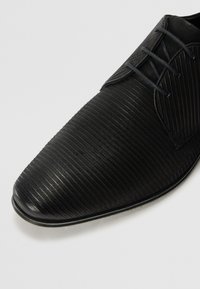 Chaussure noire avec une surface texturée et nervurée. Dotée d'un bout pointu, d'une semelle plate et de lacets noirs, mettant en avant un design épuré.