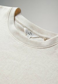Beige Rundhals-Sweatshirt mit einer weichen Textur, das ein dezentes Melange-Muster aufweist und ein Etikett mit der Aufschrift "THE SET" im Nacken hat. Größe M.
