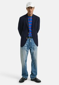 Marineblazer met gouden knopen boven een blauw-rood gestreept shirt, lichtblauwe jeans en zwarte leren schoenen, afgewerkt met een witte pet.