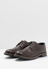 Une paire de chaussures de ville en cuir marron foncé à lacets avec des détails cousus et un talon bas empilé sur une surface blanche.