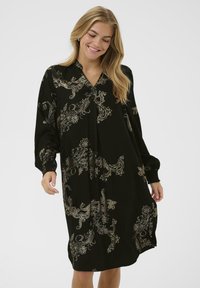 Smiješna žena s plavom kosom u crnoj dugoj haljini s zlatnim cvjetnim paisley uzorcima na svijetloj pozadini.