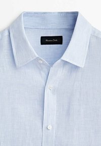 Chemise boutonnée bleu clair à fines rayures avec un col classique et une étiquette de marque noire à l'intérieur du col.