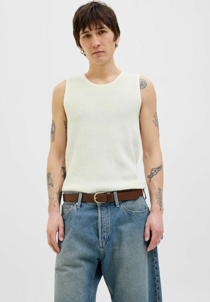 Witte geribbelde mouwloze top met ronde hals, gecombineerd met lichtblauwe losse jeans en een bruine riem, met een gestructureerd oppervlak en zichtbare tatoeages.