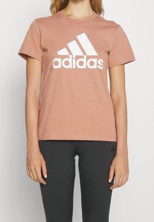 Osoba nosząca pyłowo różową koszulkę Adidas z dużym białym logo, zestawioną z ciemnoszarymi legginsami na jednolitym tle.