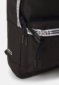 Czarny plecak z trwałego materiału, z białym logo "LACOSTE", teksturowaną powierzchnią oraz czarnymi elementami metalowymi na zapięciach. Logo Lacoste z boku.