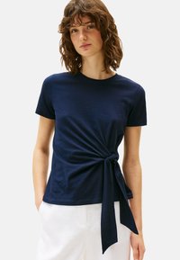 Camiseta azul marino de tela suave, con escote redondo y un detalle de nudo retorcido en la cintura, combinada con pantalones blancos.