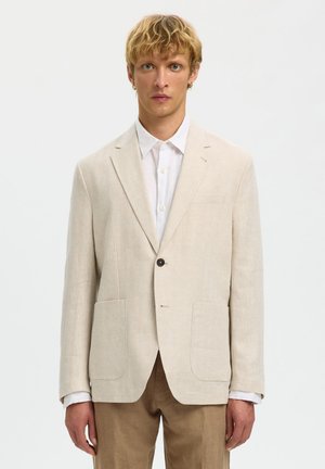 Giovane uomo con capelli biondi corti che indossa un blazer beige sopra una camicia bianca e pantaloni marrone chiaro su uno sfondo semplice.