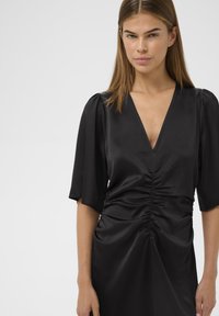 Must mustriga mustriga, millel on sügav V-kaelus, lühikesed puffvarrukad ja rükki detailid vöökoht ja seelik, valmistatud tekstureeritud kangast.