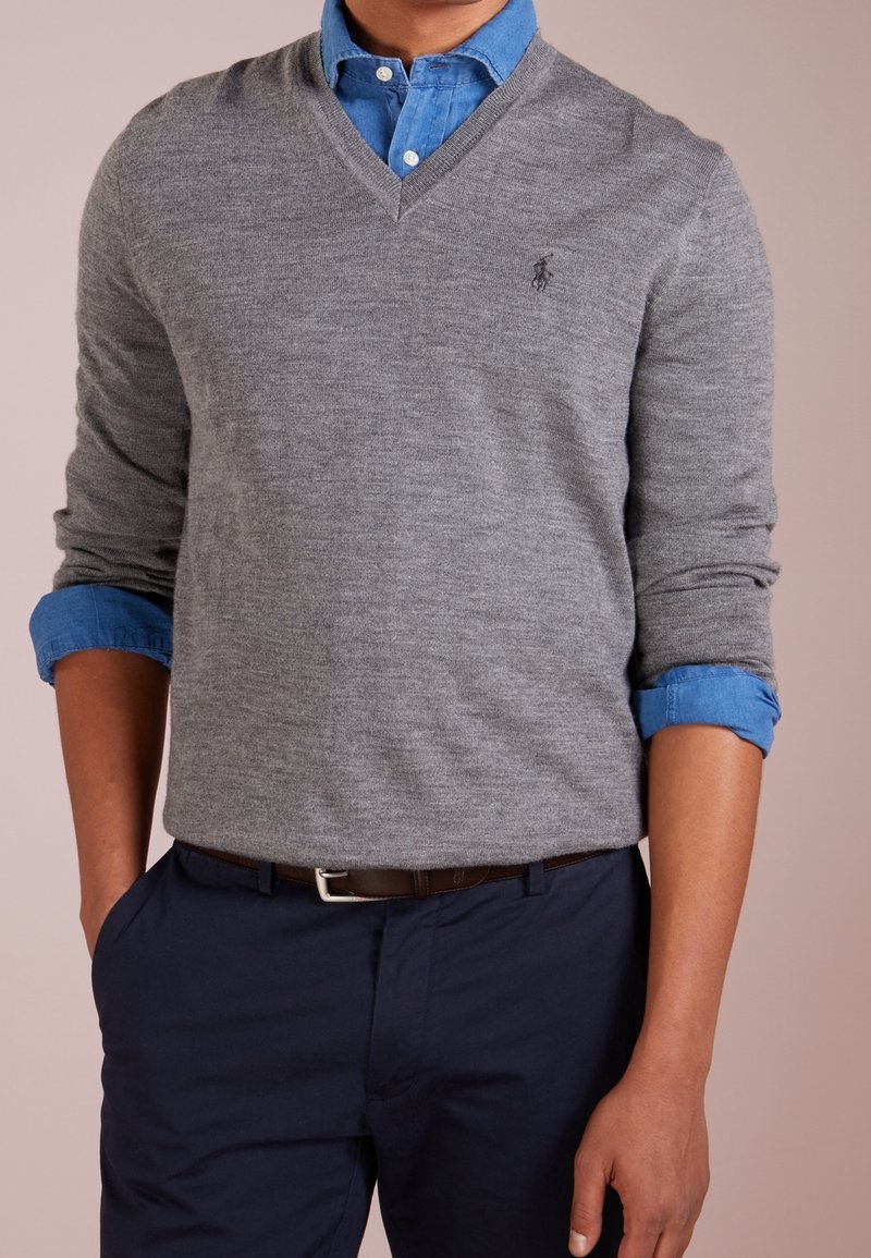 Homme portant un pull gris à col en V sur une chemise bleue à boutons avec les manches retroussées, pantalon bleu marine et ceinture marron, debout avec une main dans la poche.