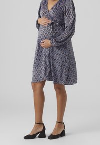 Graviditetsklänning med ett blått och brunt mönster, långa ärmar, V-ringning och knytning fram. Bärs med svarta blockklackskor.