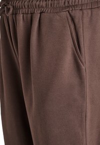 Hnedé bavlnené tepláky s elastickým pásom a šikmými bočnými vreckami, zobrazené v detailnom zábere.