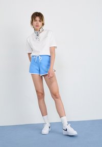 Weißes T-Shirt mit grünem Logo, blaue Sportshorts mit weißem Saum und weiße Turnschuhe, gestylt mit einem schwarz-weißen gemusterten Schal.